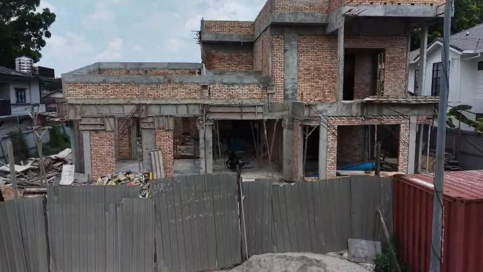 An elevated view of a two-storey house at structural stage: a reinforced-concrete frame with red clay brick infill walls, unglazed openings, and steel props and timber staging inside, behind a grey metal site hoarding.